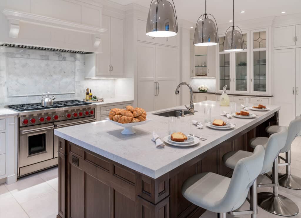 Modern kitchen in a stately home with a large island, three gray chairs, pendant lights, a gas stove, and plates with croissants and drinks on the counter.