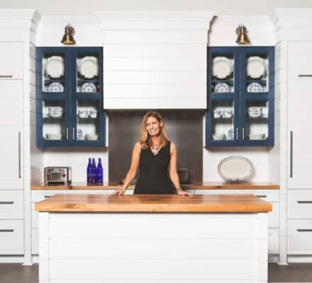 A woman stands behind a kitchen island with wood countertop, surrounded by white cabinets, blue glass-front cupboards displaying dishes, and a sleek Wolf appliance—making the space feel like a stylish kitchen showroom.