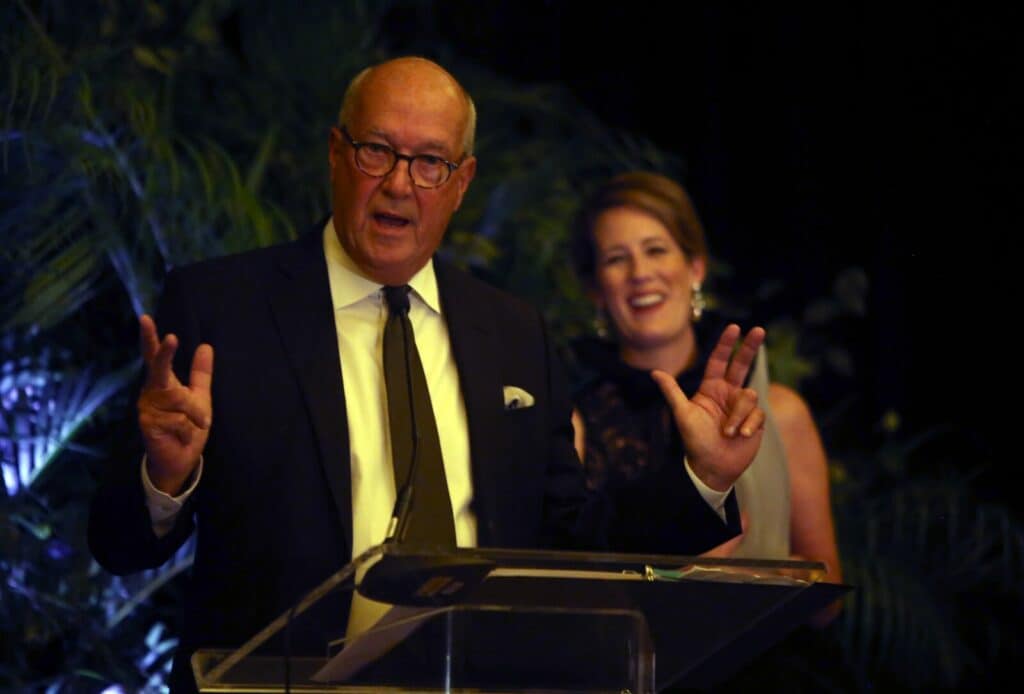 A man in a suit speaks at a podium, gesturing with both hands, while a woman in formal attire stands smiling in the background—much like unveiling an innovative Sub-Zero appliance at a prestigious event.