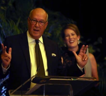 A man in a suit speaks at a podium, gesturing with both hands, while a woman in formal attire stands smiling in the background—much like unveiling an innovative Sub-Zero appliance at a prestigious event.