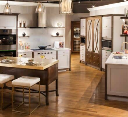 Modern kitchen with white cabinets, wooden accents, island seating, built-in Sub Zero appliance, and pendant lighting on a hardwood floor. Two place settings and glasses of white wine are set on the island.