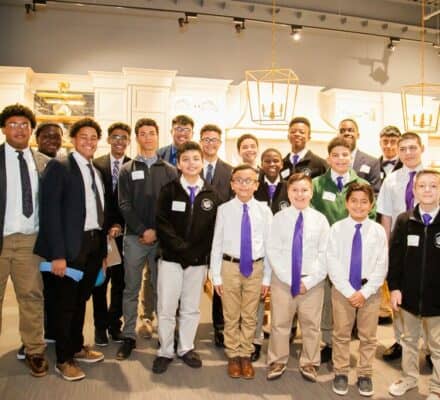 A group of boys and teenagers, dressed in semi-formal attire with ties and jackets, pose for a group photo indoors under bright lighting in a modern appliance showroom.