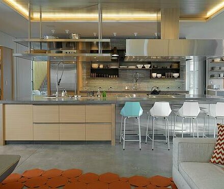 Modern kitchen with light wood cabinets, stainless steel appliances, open shelves, a large island, and five white bar stools (one blue)—a space that rivals any luxury kitchen appliance showroom.