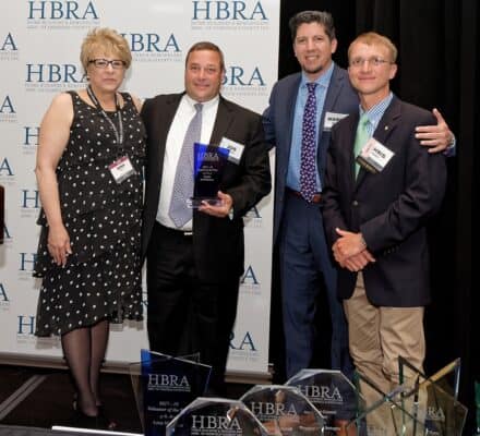 Four people stand together at an HBRA event, with one man holding a luxury kitchen appliance award. Several other awards are displayed on a table in front of them.