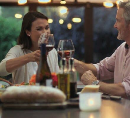 Two people sit at a table, smiling and clinking wine glasses, with food and a candle in the foreground—set in a kitchen showroom featuring the latest Wolf appliance.