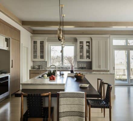 Modern kitchen showroom featuring wood and white cabinetry, a large island with barstools, pendant lights, built-in Sub Zero and Wolf appliances, plus floor-to-ceiling windows pouring in natural light.