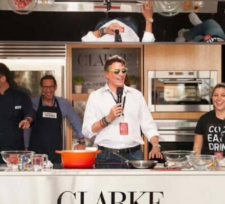 Four people stand behind a kitchen counter at a live cooking demonstration in a kitchen showroom, with luxury kitchen appliances and ingredients laid out in front of them. A mirror above reflects the bustling scene.