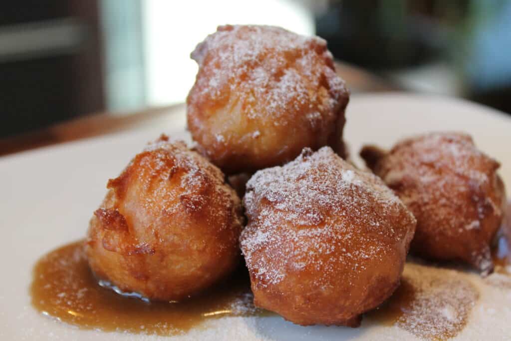 Four golden-brown fritters dusted with powdered sugar and drizzled with syrup are artfully arranged on a white plate—perfectly prepared using a luxury kitchen appliance.