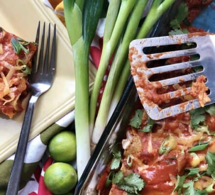 A close-up of enchiladas topped with cheese and sauce, a metal spatula, a fork, green onions, cilantro, and whole limes on a colorful tablecloth in a setting inspired by a luxury kitchen appliance showroom.
