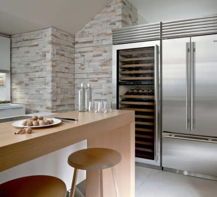Modern kitchen with light wood accents, a plate of nuts on the counter, two stools, and a Sub Zero appliance suite, including a stainless steel refrigerator and wine cooler set against a stone-tiled wall.