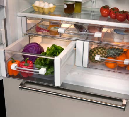 A refrigerator interior, like one in a kitchen showroom, displays eggs, tomatoes, mustard, condiments, and drawers filled with bell peppers, lettuce, cabbage, pineapple, and other fresh produce.