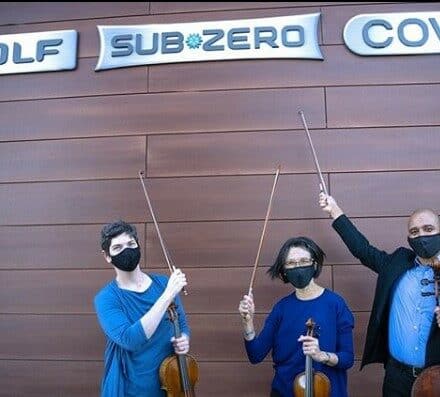 Three masked musicians holding violins and bows stand in front of a wall with Wolf and Sub-Zero appliance showroom signs.