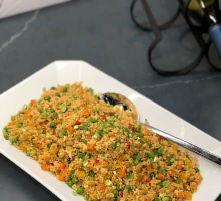 A rectangular white platter with a serving spoon displays quinoa mixed with peas, diced carrots, and red bell peppers. Two wine bottles sit in the background of a modern kitchen showroom featuring luxury kitchen appliances.