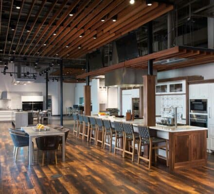 Modern, spacious kitchen with wooden flooring, a large island with bar stools, a dining table, Sub Zero appliance, stainless steel finishes, and an open ceiling with exposed beams and lighting—perfect for a kitchen showroom.