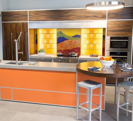 Modern kitchen with orange and wood accents, Sub-Zero and Wolf appliances, a round dining area, and a colorful backsplash depicting a landscape—perfect inspiration for any kitchen showroom.
