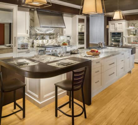 Modern kitchen with white cabinets, marble countertops, stainless steel appliances—including a Wolf appliance—island seating for two, and pendant lighting on a light wood floor.