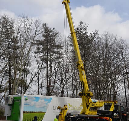 A yellow mobile crane is positioned next to a truck with a green and white container, possibly delivering a luxury kitchen appliance, while two workers stand nearby in a parking lot with trees in the background.
