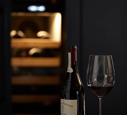 A bottle of red wine, a filled glass, a cork, and a corkscrew rest on marble, with a Sub-Zero appliance wine fridge in the dimly lit background—perfect for an elegant night in or an upscale appliance showroom display.