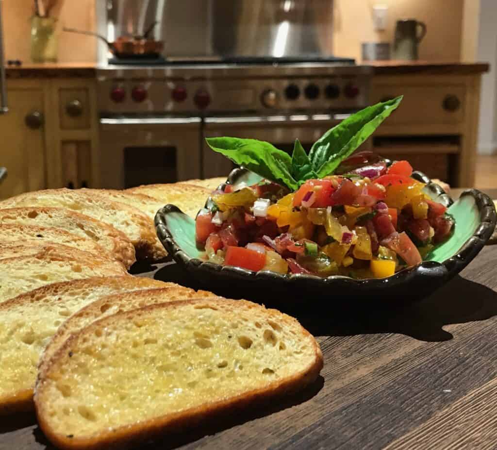 Sliced toasted bread arranged beside a bowl of tomato bruschetta topped with fresh basil, set on a wooden surface in a kitchen inspired by a luxury appliance showroom.