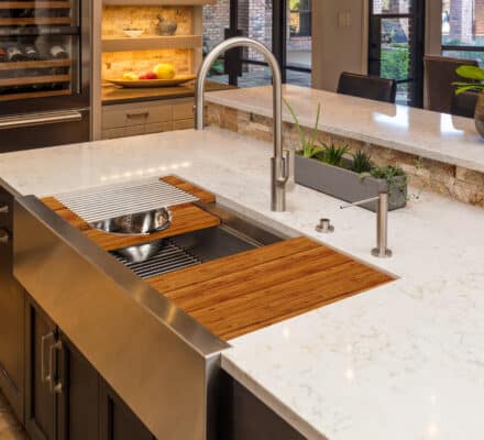 Modern kitchen island with a stainless steel sink, bamboo cutting boards, white quartz countertop, potted plant, luxury kitchen appliance, and dining area in the background.