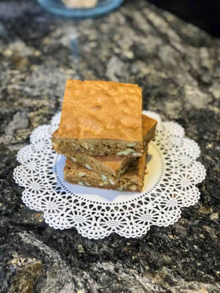 Three square blondie bars stacked on a white lace doily, placed on a dark marbled countertop, evoke the elegance of a kitchen showroom featuring premium Sub Zero appliances.