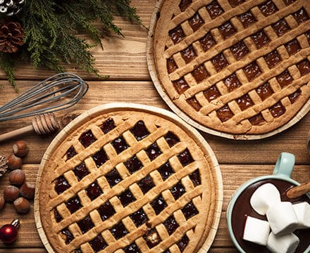 Two lattice-topped pies sit on a wooden table surrounded by a whisk, wooden spoon, holiday decorations, hazelnuts, and a cup of hot chocolate with marshmallows—perfect inspiration for your kitchen showroom or next wolf appliance creation.