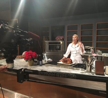 A chef in a white coat presents a cooked roast on a kitchen showroom set featuring Wolf appliances while being filmed by a camera crew.