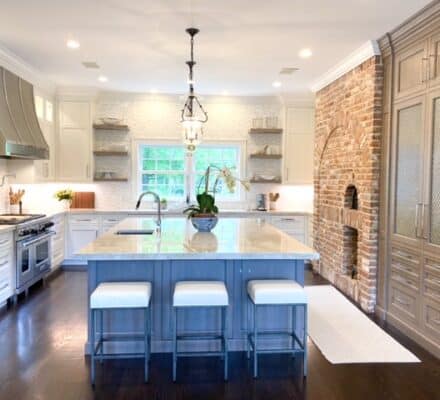 Modern kitchen with a large island, three stools, stainless steel Wolf and Sub-Zero appliances, open shelves, brick accent wall, and floor-to-ceiling cabinets—like an upscale appliance showroom.