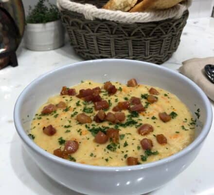 A bowl of creamy soup topped with diced ham and chopped herbs sits on a countertop, with a basket of bread in the background—an inviting scene worthy of any luxury kitchen appliance display.