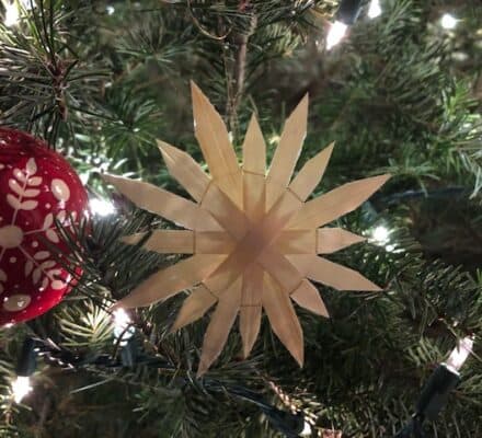A straw star ornament and a red ball with white leaf patterns hang on a lit Christmas tree, bringing festive charm to any luxury kitchen appliance showroom.