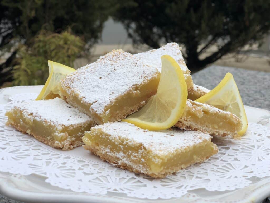 A plate of powdered sugar-dusted lemon bars is arranged with lemon slices on a white lace doily, set outdoors with greenery in the background—perfect inspiration for your next visit to a kitchen showroom.