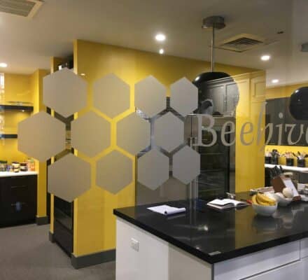 Modern office kitchen with yellow walls, black countertops, and "Beehive" text with hexagonal designs on a glass partition; reminiscent of a stylish kitchen showroom, various kitchen items are visible.