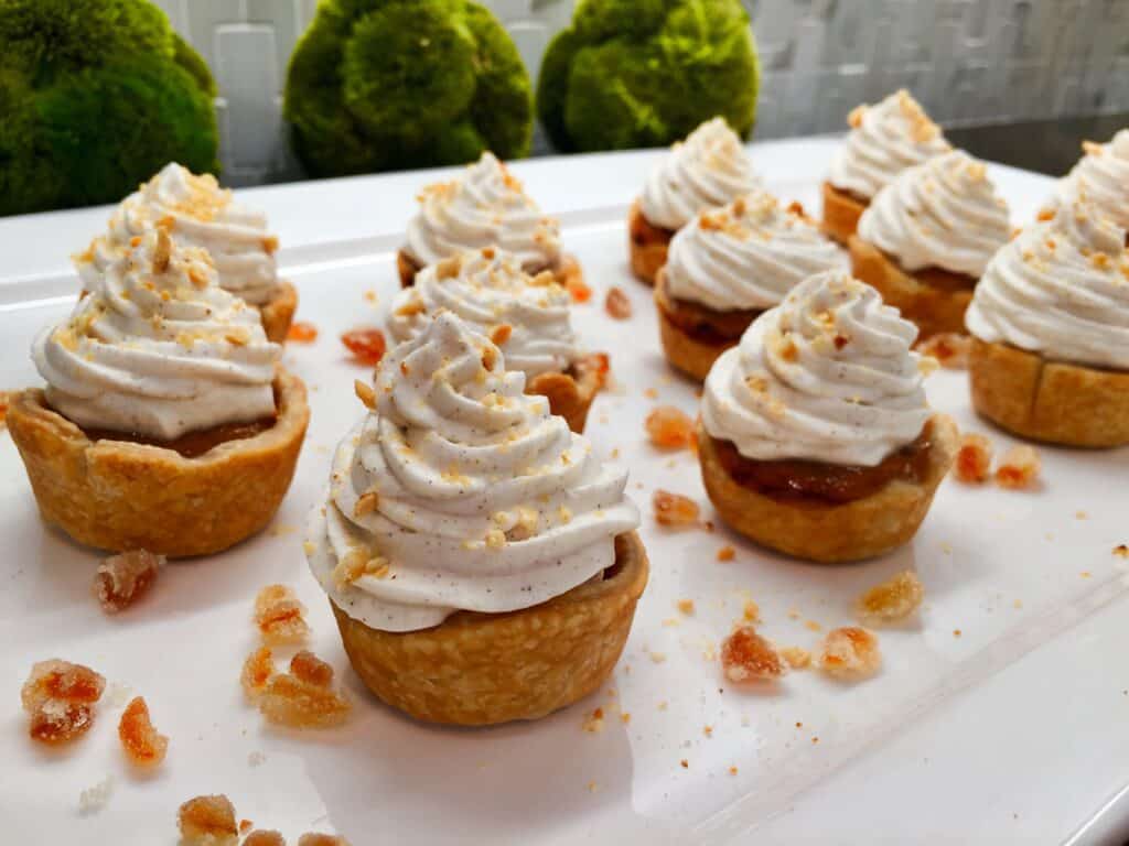 Mini tarts with swirled whipped topping, sprinkled with crushed nuts, are arranged on a white platter with caramelized sugar pieces—ideal for serving in an appliance showroom featuring the latest luxury kitchen appliances. Green moss decor is visible in the background.