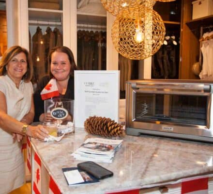 Two women stand at a kitchen showroom counter beside a large toaster oven, pamphlets, and decorations, including a Canadian flag and a pinecone.