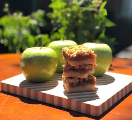 Three green apples and a stack of three crumbly dessert bars rest on a striped cutting board, with sunlight and greenery in the background—an inviting scene reminiscent of a luxury kitchen appliance showroom.
