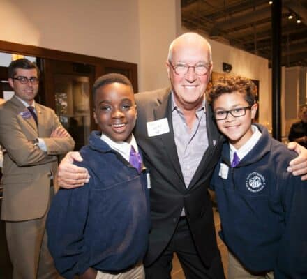 An older man with glasses stands smiling between two boys in school uniforms at an indoor event, possibly a kitchen showroom, with another man standing in the background.