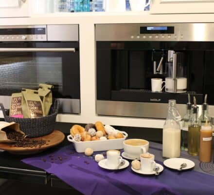 A kitchen showroom counter with a built-in coffee machine, two cups, bottles of syrup, a bowl of coffee beans, a basket of pastries, and coffee drinks on a purple cloth displays the perfect luxury kitchen appliance setup.