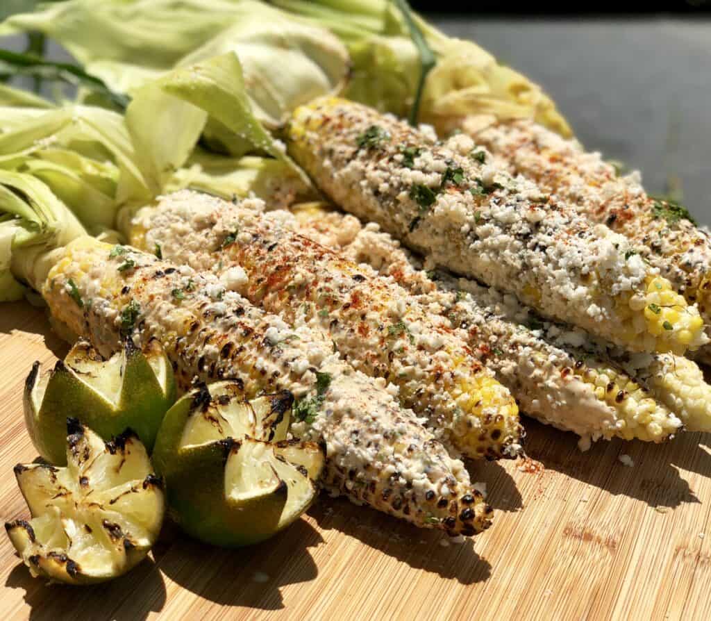 Grilled corn on the cob topped with cheese and spices, prepared using a Wolf appliance, and served on a wooden board with charred lime wedges.
