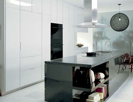 Modern kitchen showroom featuring white cabinets, a black island countertop, built-in Sub-Zero appliance, open shelving, a round light fixture, and a dining area with wooden chairs and a nature-themed wall mural.