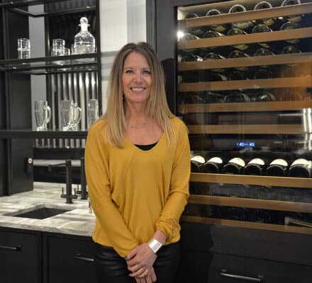 A woman wearing a yellow top stands in a modern kitchen showroom next to a wine fridge filled with bottles and glassware displayed on shelves.