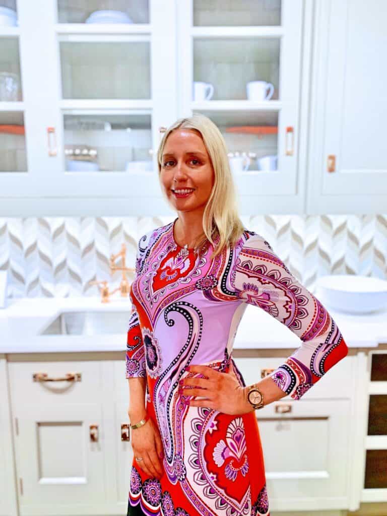A woman with blonde hair wearing a patterned dress stands in a modern kitchen showroom with white cabinets, a chevron backsplash, and luxury kitchen appliances.