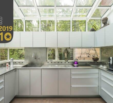 Modern kitchen with light grey cabinets, stainless steel countertops, a glass ceiling, and luxury kitchen appliances—including coffee machines and decorative objects; “Top Ten Monthly Gold 2019” badge in the corner.