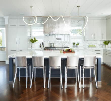 Modern kitchen with white cabinets, marble countertops, luxury kitchen appliances, an island with five metal chairs, and a sculptural light fixture overhead.