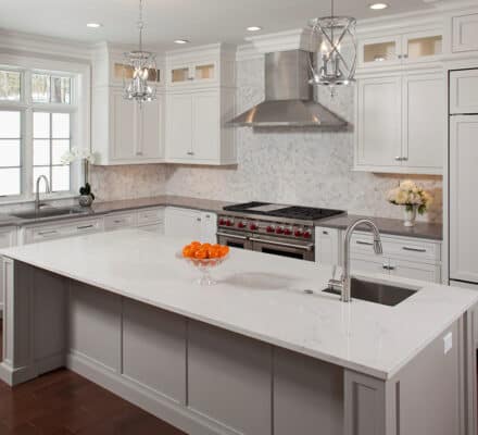 Modern kitchen with white cabinets, stainless steel appliances including a Wolf appliance, a marble island with a sink, and a bowl of oranges as centerpiece under pendant lights.