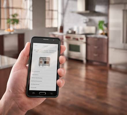 A person holds a smartphone displaying appliance information in a modern kitchen with stainless steel appliances, wooden cabinets, and a luxury kitchen appliance reminiscent of a high-end kitchen showroom.