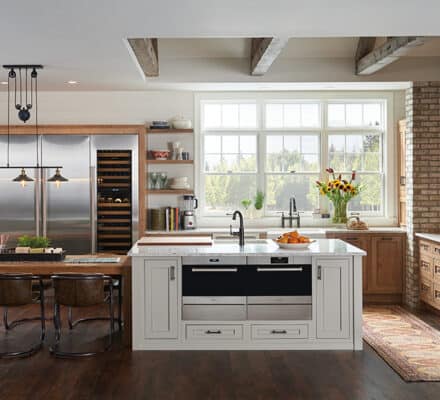 Modern kitchen showroom featuring exposed brick walls, wooden cabinetry, a central island, stainless steel and Sub-Zero appliances, a large window, and a dining table with four chairs.