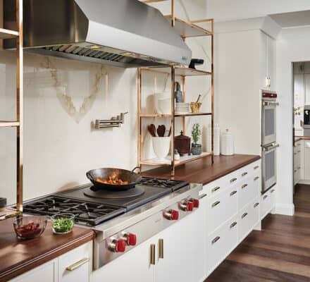 Modern kitchen with white cabinets, wooden countertops, open shelving, and a gas stove with a wok. Stainless steel appliances, including a Wolf appliance, and dark wood flooring create an elegant look.
