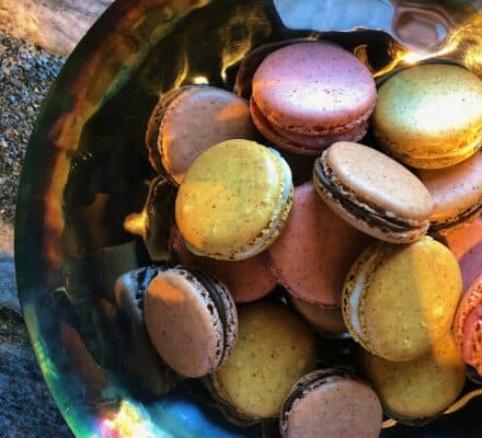 A metallic bowl filled with assorted colorful macarons sits on a stone surface in a kitchen showroom, illuminated by natural light.