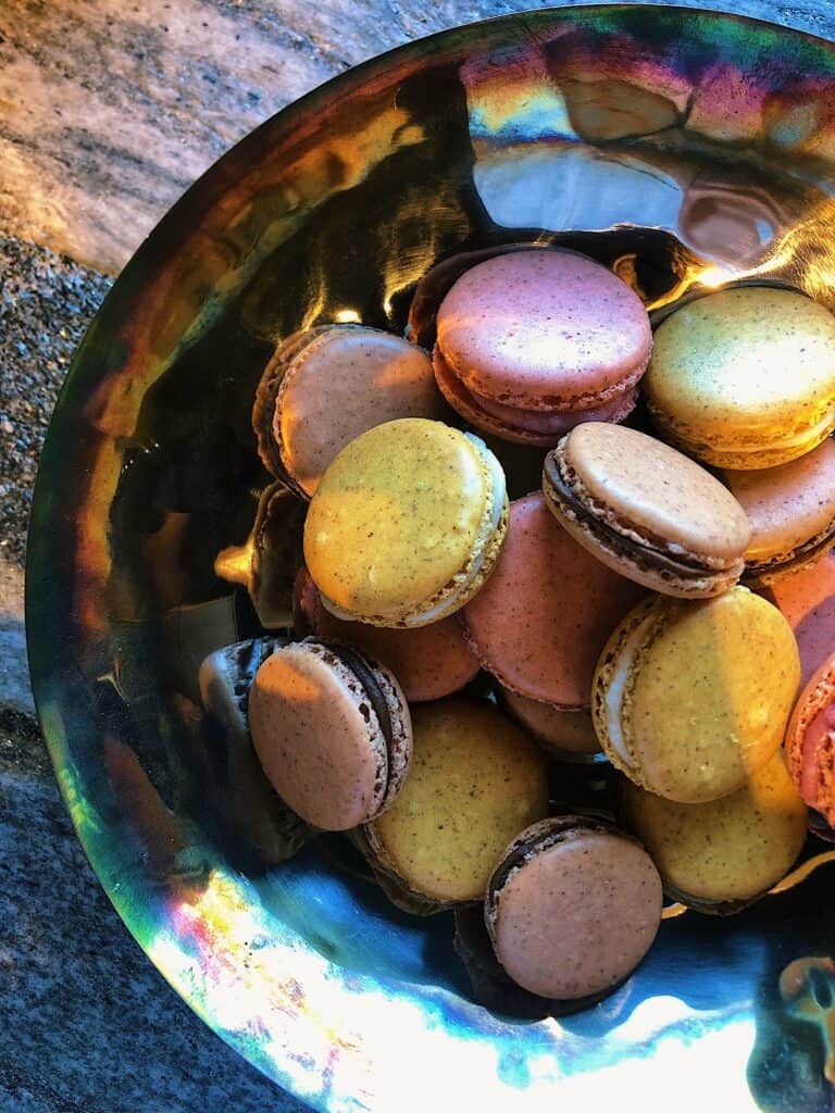 A metallic bowl filled with assorted colorful macarons sits on a stone surface in a kitchen showroom, illuminated by natural light.