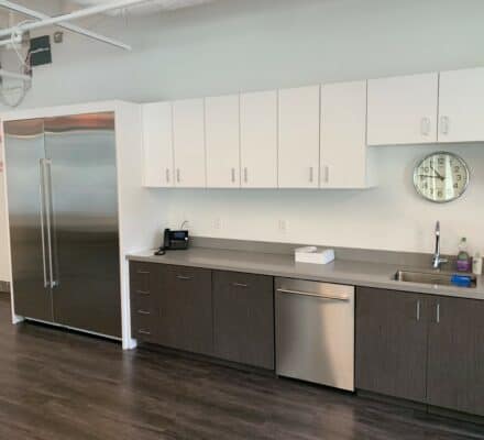 Modern office kitchen showroom featuring white upper cabinets, dark lower cabinets, Wolf and Sub-Zero appliances, a sink, phone, wall clock, and soap dispenser on a sleek gray countertop.